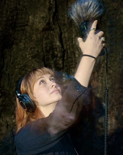 Headshot of Raegan Padula; she is looking up and holding an audio recording device while wearing headphones